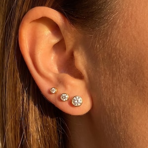 Peut inclure: Trois boucles d'oreilles à clou en or avec des pierres rondes transparentes serties dans une monture en biseau, disposées en rangée sur le lobe de l'oreille. Chaque boucle présente un bord en or poli entourant une pierre blanche étincelante.