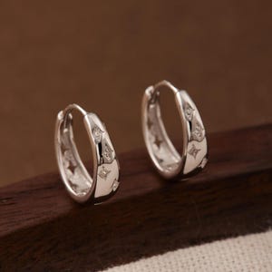 May include: A pair of silver hoop earrings featuring a polished finish and small, engraved four-pointed star motifs spaced evenly along the outer surface of each hoop. The earrings have a hinged closure mechanism and a rounded, thick profile.