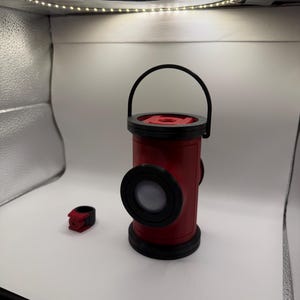 May include: A red and black cylindrical lantern featuring a circular emblem on the top and a side lens, accompanied by a small matching red and black ring-shaped accessory. The lantern has a black handle and a solid base, designed with a distinct aesthetic.