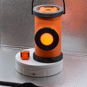 May include: A cylindrical orange lantern with a black handle and a circular glowing orange center, accompanied by a small square orange and black light-up component, both resting on a white circular rotating display stand.