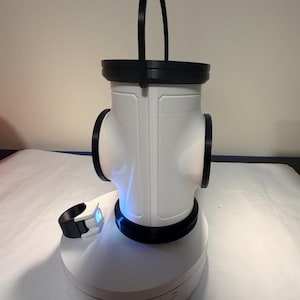 May include: A white cylindrical device with black accents and a top handle, featuring a central body with side ports. It sits on a circular white base with a small black and white component nearby that emits a blue light from its side.