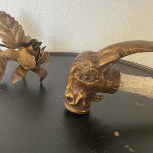 May include: A vintage-style decorative metal rose and a unique, antique-looking handle with a gold-colored ram's head. The handle is attached to a light-colored, natural-looking wooden stick. The items are gold-colored and sit on a dark surface.