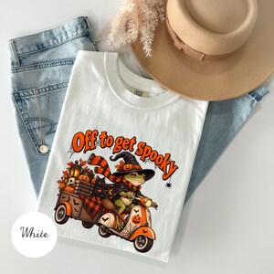 Op de afbeelding: Een wit t-shirt met een afbeelding van een kikker met een heksenhoed en sjaal, rijdend op een oranje en witte scooter. De kikker trekt een karretje vol met pompoenen, een lantaarn en boeken. De tekst luidt Off to get spooky in het oranje.