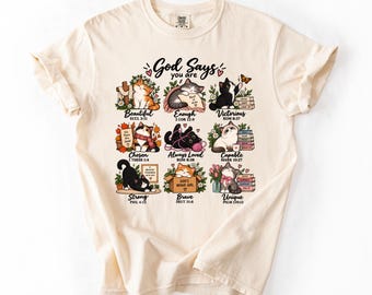 May include: An ivory t-shirt featuring a graphic print of nine illustrated cats with the text God Says you are and various inspirational phrases including Beautiful, Enough, Victorious, Chosen, Always Loved, Capable, Strong, Brave, and Unique.