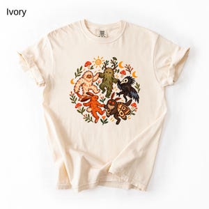 Op de afbeelding: Een ivoorkleurig t-shirt met een cirkelvormige afbeelding van mythische wezens, waaronder een yeti, een bosgeest, een vogel als pestmeester, een konijn en een mot die samen dansen, omringd door kleine paddenstoelen, bladeren, een zon en een maansikkel.