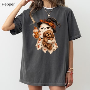 Puede incluir: Una camiseta gris carbón con un gráfico de un fantasma con sombrero de bruja, sosteniendo una taza de café con temática de calabaza y un cachorro marrón. El diseño incluye calabazas naranjas, hojas de otoño y lunas en el cuerpo del fantasma.
