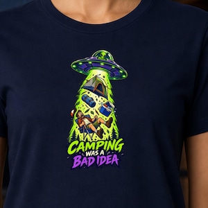 May include: A navy blue short-sleeve t-shirt featuring a graphic of a green UFO abducting a camper, a tent, and gear. The design includes the text "CAMPING WAS A BAD IDEA" in bright green and purple lettering centered on the chest of the garment.
