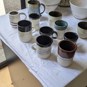 May include: A collection of ceramic mugs and bowls featuring various glaze colours including white, black, brown, and blue. The mugs have cylindrical shapes with handles, while the bowls are wide and open. Some pieces display horizontal line patterns.