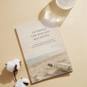 Può includere: Un quaderno rettangolare con copertina beige che riporta il testo LE PAROLE CHE NON HAI MAI DETTO e una fotografia di un messaggio in una bottiglia su una spiaggia sabbiosa. Un rametto di cotone bianco è posizionato accanto al quaderno.