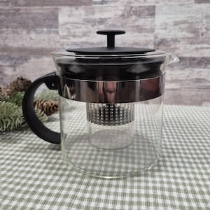 May include: A clear glass teapot with a black handle, lid, and infuser. The teapot has a stainless steel band around the middle. The background is a light wood-grain pattern and a green and white checkered tablecloth.