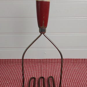 Puede incluir: Antiguo pasapurés con mango de madera rojo y blanco y cabezal de acero inoxidable. El mango tiene un aspecto desgastado. El pasapurés está sobre un mantel a cuadros rojos y blancos.