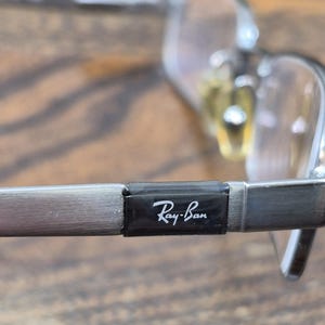 K&ouml;nnte beinhalten: Silberfarbene Metallbrille mit einem rechteckigen schwarzen B&uuml;geleinsatz, auf dem das wei&szlig;e Ray-Ban-Logo aufgedruckt ist. Der Rahmen verf&uuml;gt &uuml;ber klare Gl&auml;ser und verstellbare Nasenpads und zeigt ein metallisches Finish.