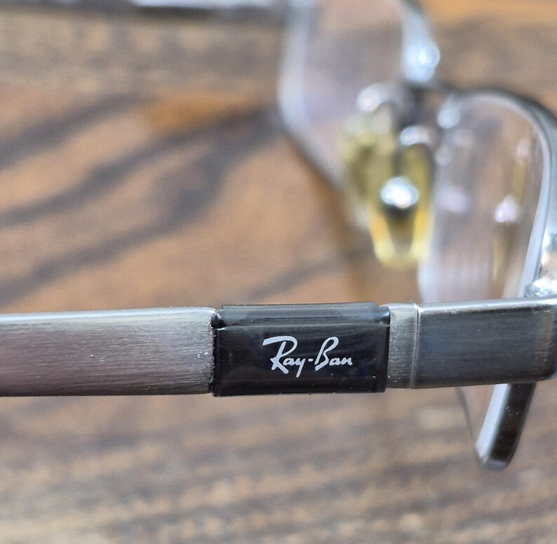 K&ouml;nnte beinhalten: Silberfarbene Metallbrille mit einem rechteckigen schwarzen B&uuml;geleinsatz, auf dem das wei&szlig;e Ray-Ban-Logo aufgedruckt ist. Der Rahmen verf&uuml;gt &uuml;ber klare Gl&auml;ser und verstellbare Nasenpads und zeigt ein metallisches Finish.