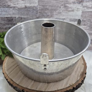 Puede incluir: Un molde para bizcocho vintage de aluminio con un tubo central y dos asas. El molde circular se asienta sobre una rodaja de madera. El molde está diseñado para hornear pasteles y tiene un acabado plateado.