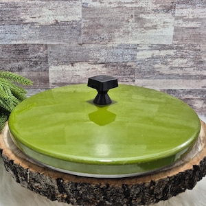 Könnte beinhalten: Ein runder, limettengrüner Keramikdeckel mit einem schwarzen quadratischen Griff. Der Deckel steht auf einem rustikalen Holzsockel, mit einem sichtbaren weißen Rand. Der Hintergrund zeigt eine strukturierte, grau-braune Holzwand und eine grüne Pflanze.