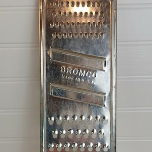 May include: A vintage metal grater with a rectangular grating surface and a black metal handle. The grater has the brand name "BROMCO" and "MADE IN U.S.A." stamped on the surface. The grater is approximately 12 inches long.