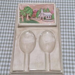 May include: A beige ceramic spoon rest with two spoon-shaped indentations. The top features a raised relief scene of a church with trees and a mountain in the background. The spoon rest is rectangular with a raised border.