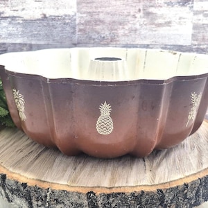 May include: A brown and cream-colored bundt pan with a pineapple design. The pan is sitting on a wooden slice and has a fluted design. The pineapple design is repeated around the pan.