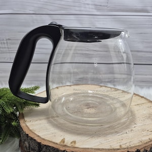 May include: A clear glass coffee carafe with a black handle and lid. The carafe has measurement markings and a warning label. The carafe is sitting on a wooden surface.