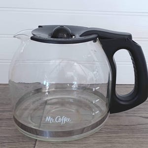 Puede incluir: Una jarra de café transparente de la marca Mr. Coffee con asa, tapa y borde negros. La jarra tiene marcas de medición y una banda plateada alrededor de la base. Está diseñada para contener hasta 12 tazas de café.
