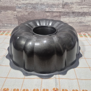 May include: A dark gray, fluted bundt pan with a central hole. The pan has a scalloped base and a metallic finish. It sits on a checkered tablecloth, ready for baking. The pan is designed for making cakes and other baked goods.
