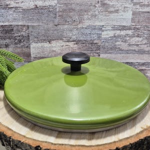 Könnte beinhalten: Ein runder, limettengrüner Keramikdeckel mit schwarzem Knopf. Der Deckel liegt auf einer Holzscheibe mit Rindenkante. Der Hintergrund ist eine Holzdielenwand. Eine grüne Pflanze befindet sich in der linken Ecke.