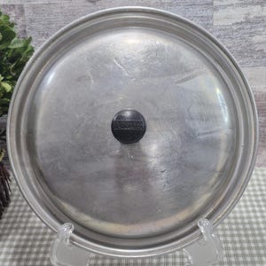 Puede incluir: Tapa redonda de metal plateado con un pomo negro. El pomo tiene la inscripción "WEAREVER". La tapa está ligeramente rayada y muestra signos de uso. Probablemente una tapa para una olla o sartén.