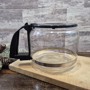 May include: A clear glass coffee carafe featuring a black plastic handle and a matching black lid. The carafe has a silver-toned metal band around the base and includes measurement markings on the side, with the text MR. COFFEE visible on the glass.