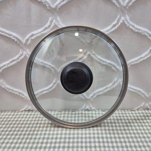 May include: A circular, transparent glass pot lid featuring a stainless steel rim and a central black knob. The word Tramontina is embossed on the black knob. The lid is designed for cookware and provides a clear view of the contents inside.