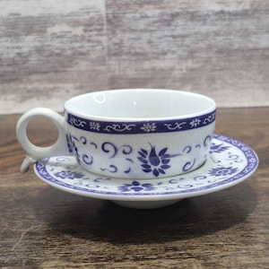 May include: White teacup and saucer set with blue floral and scrollwork design. The teacup has a handle and the saucer is round. The set is made of ceramic and is placed on a wooden surface.
