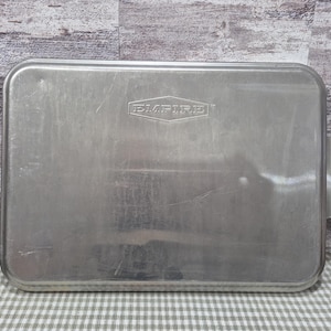 May include: A rectangular, silver baking tray with the embossed word "EMPIRE" in the center. The tray has rounded corners and a handle on one side. The tray is made of metal and is designed for baking.