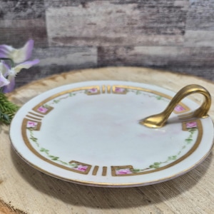 May include: A white, round, vintage porcelain serving dish with a gold handle. The rim is decorated with gold trim, pink floral accents, and green leaves. The dish is placed on a wooden surface.