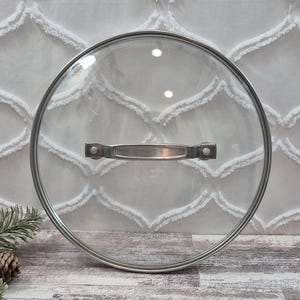 Könnte beinhalten: Ein runder Deckel aus gehärtetem Glas mit einem Rand aus poliertem Edelstahl und einem horizontalen Edelstahlgriff, der mit zwei Nieten befestigt ist. Das Glas ist transparent und das Wort essentials ist unten in kleinen Buchstaben eingraviert.