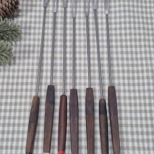 May include: Set of eight fondue forks with stainless steel tines and long, wooden handles. The handles are a dark brown color, with a variety of colored tips including red, yellow, and green. The forks are arranged on a green and white checkered tablecloth.