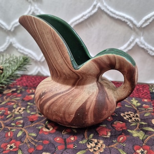 May include: A ceramic pitcher featuring a rounded base and a high, curved spout. The exterior displays a marbled pattern of light and dark brown tones, while the interior and the top edge of the handle are finished in a solid, deep forest green glaze.