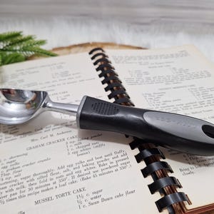 Puede incluir: Una cuchara para helado de acero inoxidable con un mango negro y gris, apoyada en un libro de cocina abierto. La cuchara tiene un cuenco redondeado y un mecanismo de palanca. Las páginas del libro de cocina están amarillentas con texto.