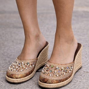 May include: A pair of embellished wedge sandals. The open-back shoes feature a woven jute wedge heel and a decorative upper with beads and pearls. The color palette includes gold, brown, and cream tones, creating a bohemian aesthetic.