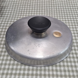 May include: A round, silver-colored lid with a black knob. The knob has the word "MICRO" printed on it. A small, gold-colored metal piece is on the lid's surface. The lid is likely for a pot or pan.