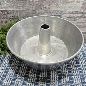 Puede incluir: Un molde redondo de aluminio para bizcocho de ángel, de época, con un tubo central. El molde tiene un cuenco ancho y poco profundo y una superficie ligeramente texturizada. El molde está sobre un paño con estampado azul y blanco.