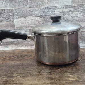 Puede incluir: Una cacerola de acero inoxidable con tapa y asa negra. La olla tiene un fondo de color cobre y un pomo negro en la tapa. El asa tiene un lazo de metal.