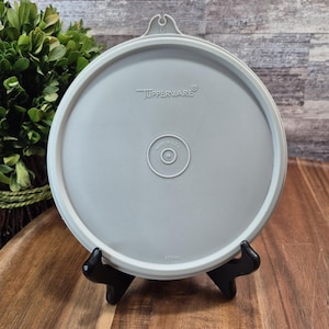 以下が含まれることがあります： ブランド名がエンボス加工された、明るい灰色のTupperwareの蓋。蓋は円形のデザインで、小さなハンドルが付いており、「Made in USA」とマークされています。黒いスタンドに展示されています。
