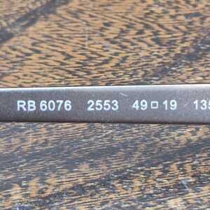 K&ouml;nnte beinhalten: Ein dunkelgrauer Brillenb&uuml;gel mit wei&szlig; aufgedrucktem Text RB 6076 2553 49 19 135. Die Oberfl&auml;che des Rahmens hat ein glattes, mattes Finish und die Beschriftung ist auf dem dunklen Hintergrund des Brillenteils deutlich zu erkennen.