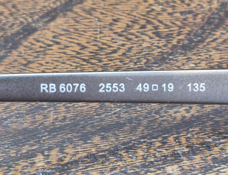 K&ouml;nnte beinhalten: Ein dunkelgrauer Brillenb&uuml;gel mit wei&szlig; aufgedrucktem Text RB 6076 2553 49 19 135. Die Oberfl&auml;che des Rahmens hat ein glattes, mattes Finish und die Beschriftung ist auf dem dunklen Hintergrund des Brillenteils deutlich zu erkennen.