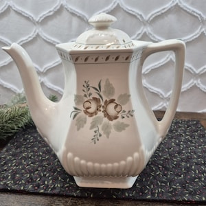 May include: A cream-colored ceramic teapot with a hexagonal body and a matching lid. The teapot features a painted floral motif in shades of brown and sage green, with a decorative leaf pattern along the upper rim and vertical fluting at the base.