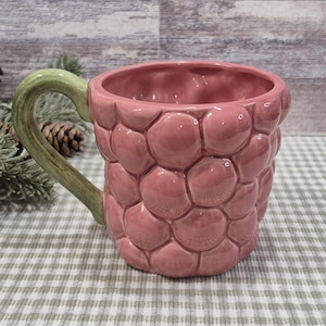 May include: A pink ceramic mug with a textured, bubble-like exterior. The handle is green and curved. The mug is designed to resemble a raspberry. The mug is on a checkered surface.