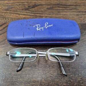 K&ouml;nnte beinhalten: Eine silberfarbene Metallbrille mit rechteckigen Gl&auml;sern und schwarzen B&uuml;gelenden, platziert vor einem blauen, rechteckigen Hartschalenetui der Marke Ray-Ban mit sichtbaren Kratzern und Gebrauchsspuren auf der Oberfl&auml;che.