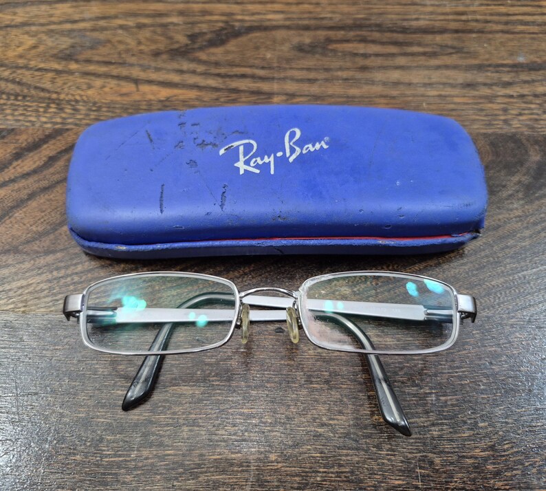 K&ouml;nnte beinhalten: Eine silberfarbene Metallbrille mit rechteckigen Gl&auml;sern und schwarzen B&uuml;gelenden, platziert vor einem blauen, rechteckigen Hartschalenetui der Marke Ray-Ban mit sichtbaren Kratzern und Gebrauchsspuren auf der Oberfl&auml;che.
