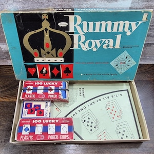 May include: An open vintage "Rummy Royal" game box with a teal lid featuring a gold crown and card suit symbols. Inside are card decks and poker chips. The box reads "a game for the whole family."