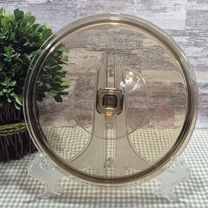 May include: A transparent, circular glass lid with a brown edge and a square handle. The lid is displayed on a clear plastic stand. The background includes a brick wall and a green plant.
