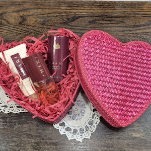 May include: A pink woven heart-shaped gift box, partially open to show beauty products. Inside are burgundy-toned items: a cream tube, a perfume bottle, and a cylindrical object, nestled on red paper. The box sits on a white lace doily.
